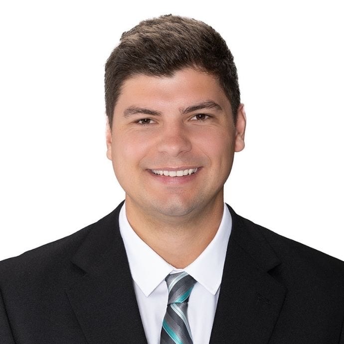 Smiling man in a dark suit, white shirt, and tie, photographed against a plain white background.