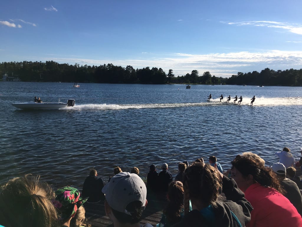 People watching a group of water skiers performing on a lake under a clear blue sky
