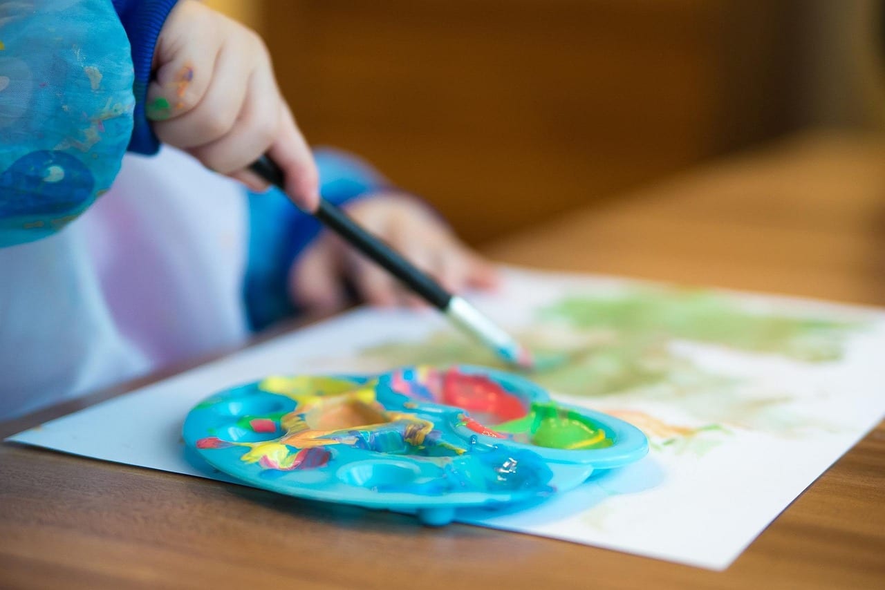 Close-up of a child's hand holding a paintbrush and painting with a colorful palette on paper.