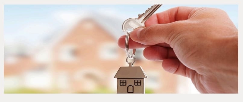 Close-up of a hand holding keys with a house-shaped keychain, blurred brick houses in the background